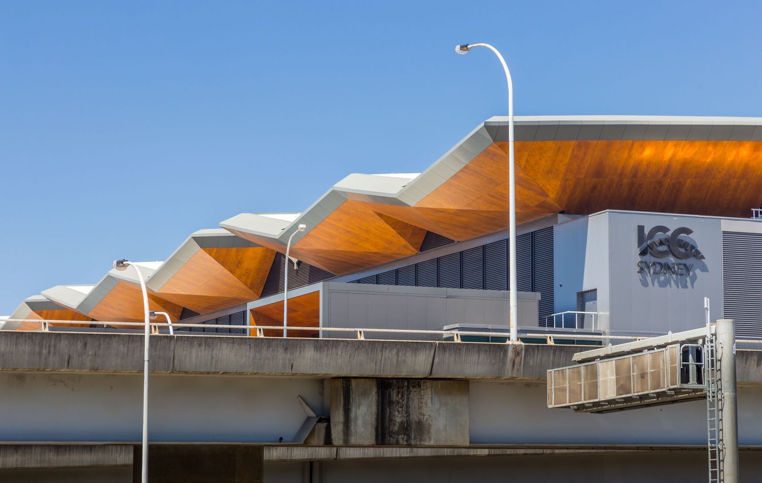 Convention centre exterior with a sculptural orange timber roofline, street lamps, and a blue sky above an elevated roadway.