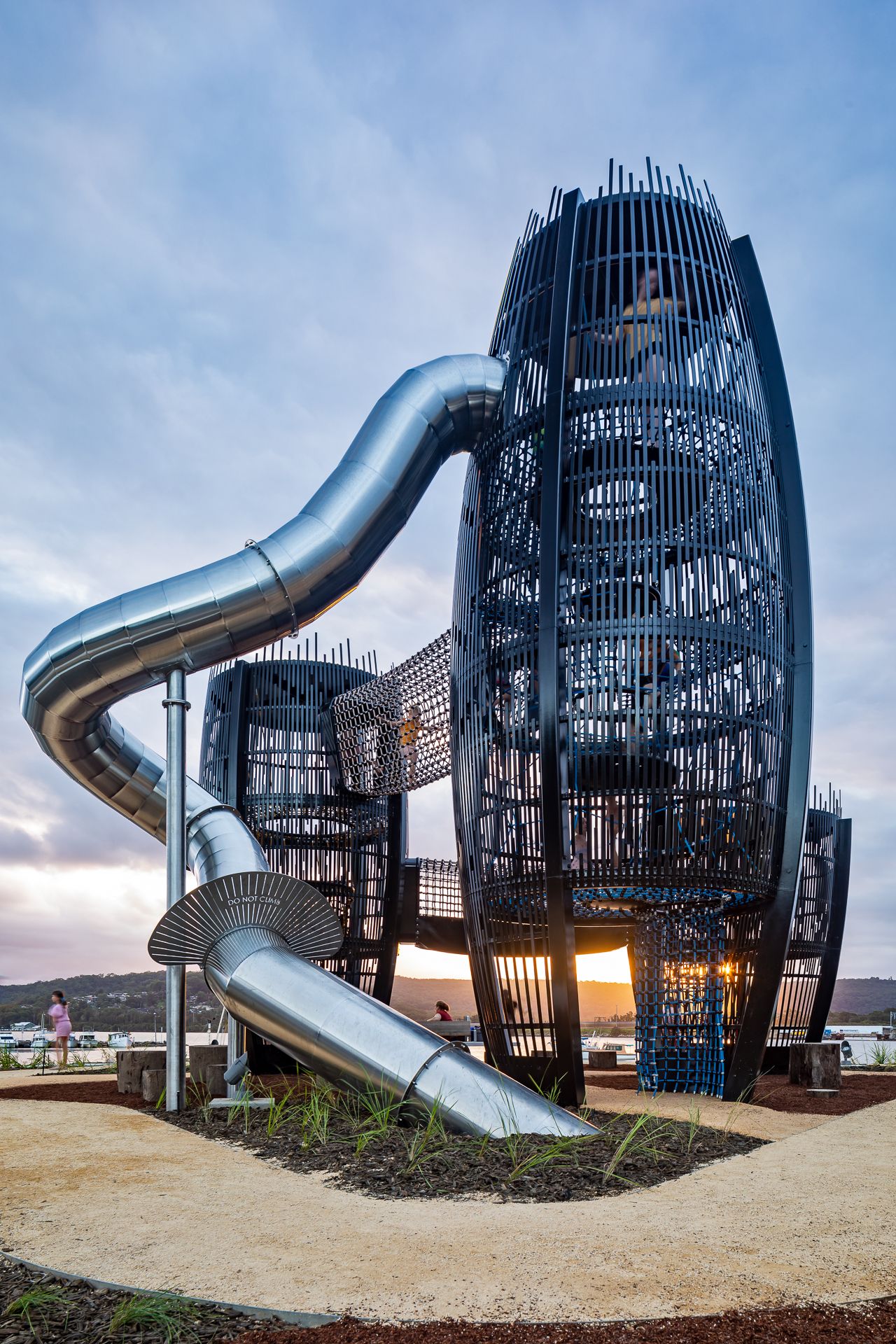 Large stainless steel tube slide spirals from a black lattice tower playground at sunset in a landscaped waterfront park.