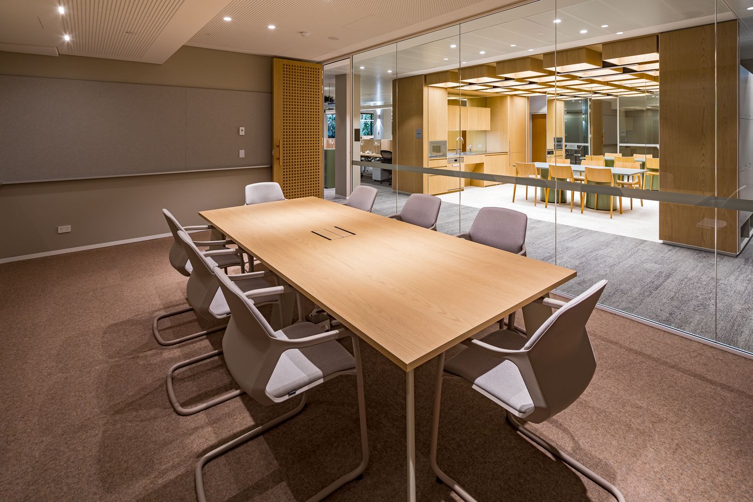 Office meeting room with a light wood conference table, gray upholstered chairs and glass walls overlooking a timber-lined kitchen.