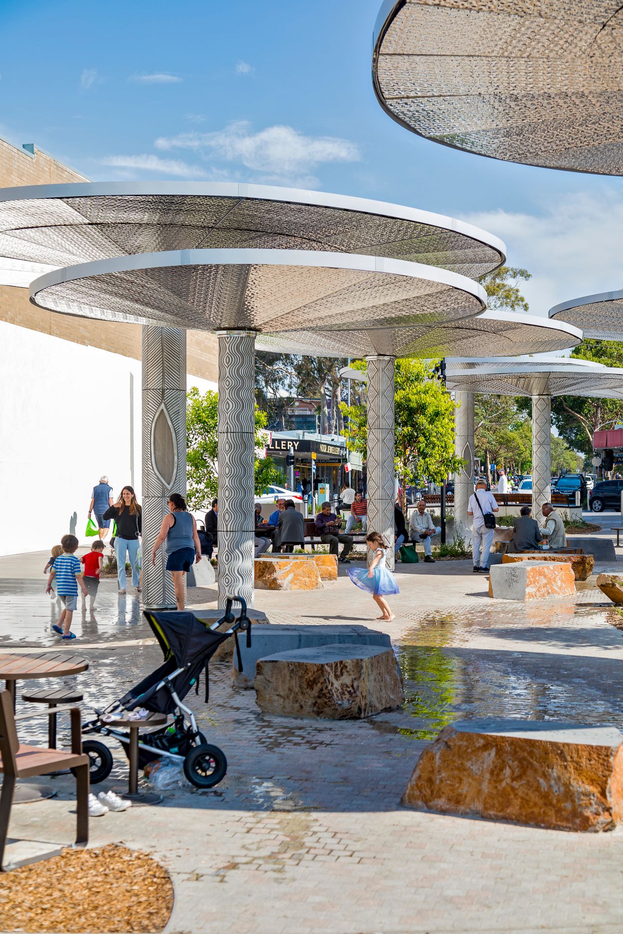 Families relax beneath large perforated metal canopies in a sunny public plaza with sandstone boulder seating and a shallow water feature.