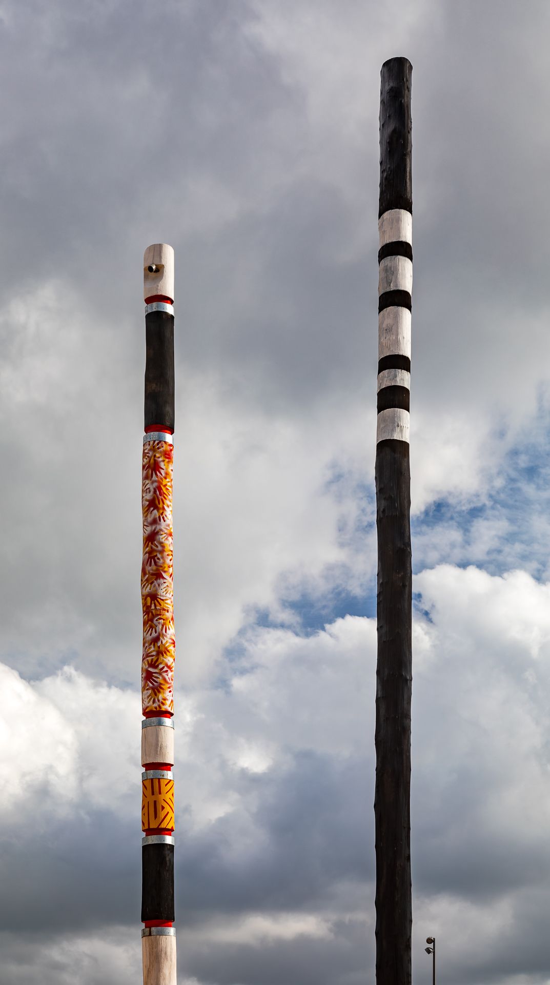 Two tall painted poles, one with orange patterns and one black with white stripes, stand against a cloudy blue sky.