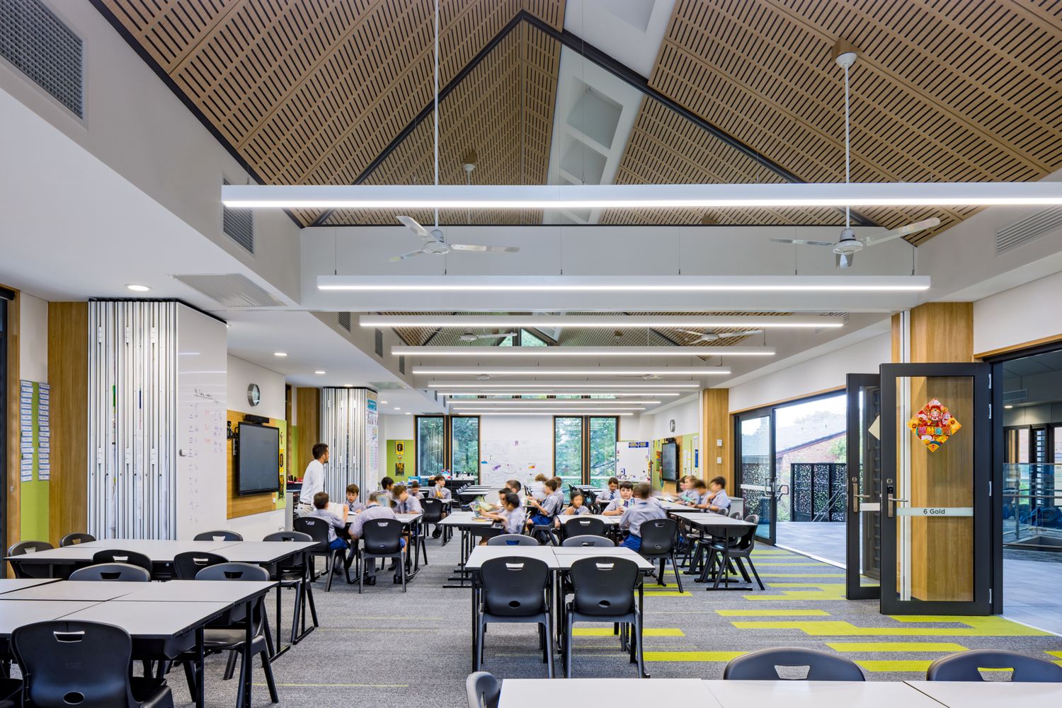 Modern school learning space with timber-slat vaulted ceiling, students seated at tables and large windows opening to outdoor areas.
