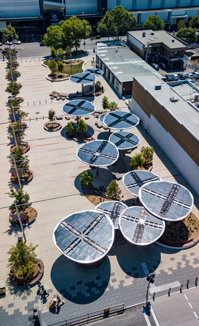 Aerial view of a paved plaza with circular solar panel canopies, planted beds and tree-lined edges beside modern buildings.