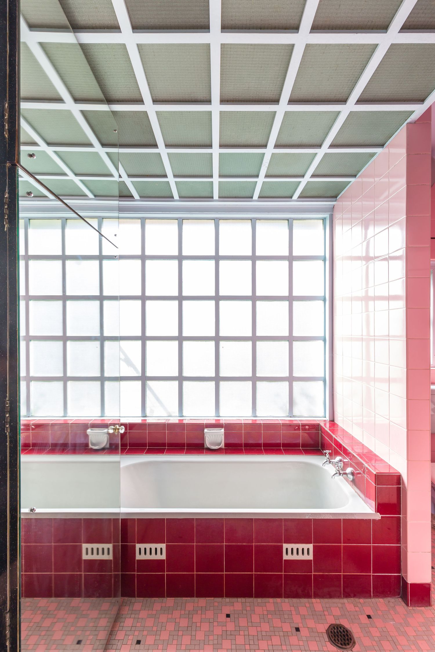 Retro bathroom with red and pink tiles, a built-in tub, a glass block window, and a grid ceiling of frosted panels.