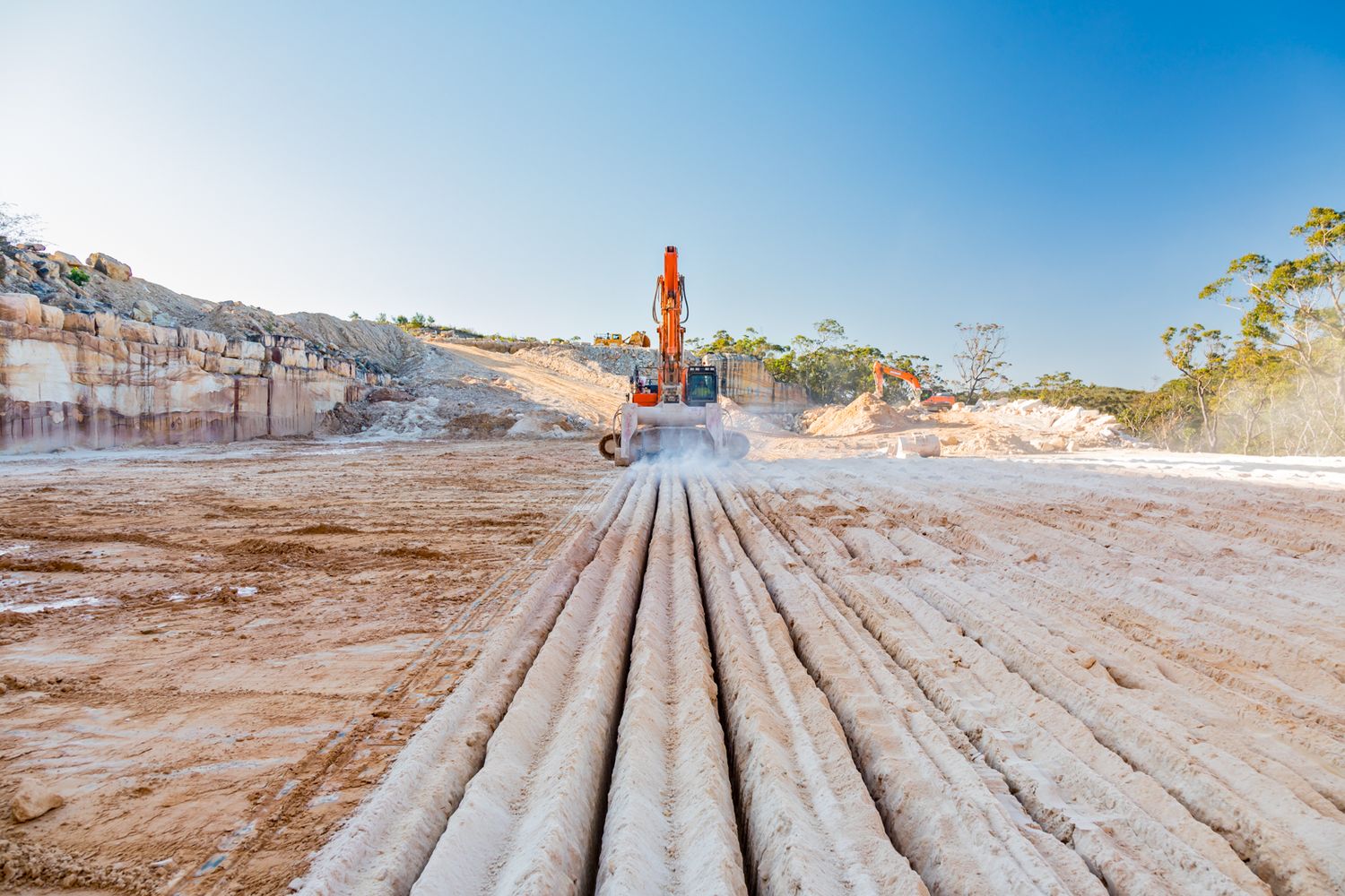 Orange excavator working at a sandstone quarry site with rows of cut stone logs in the foreground under a clear blue sky.