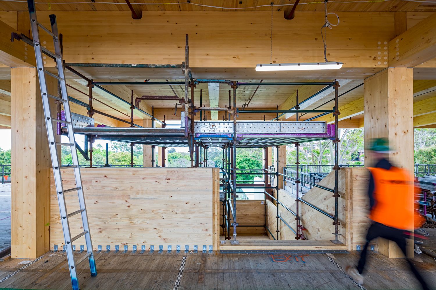 Construction worker in orange hi-vis walks past a ladder and scaffolding inside a timber-framed building under construction.