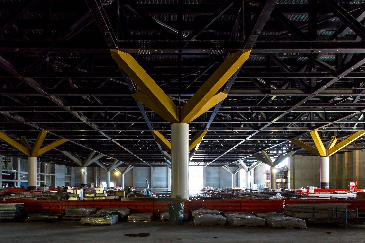 Large dark industrial interior with yellow angled steel columns supporting a high ceiling and construction materials stacked below.