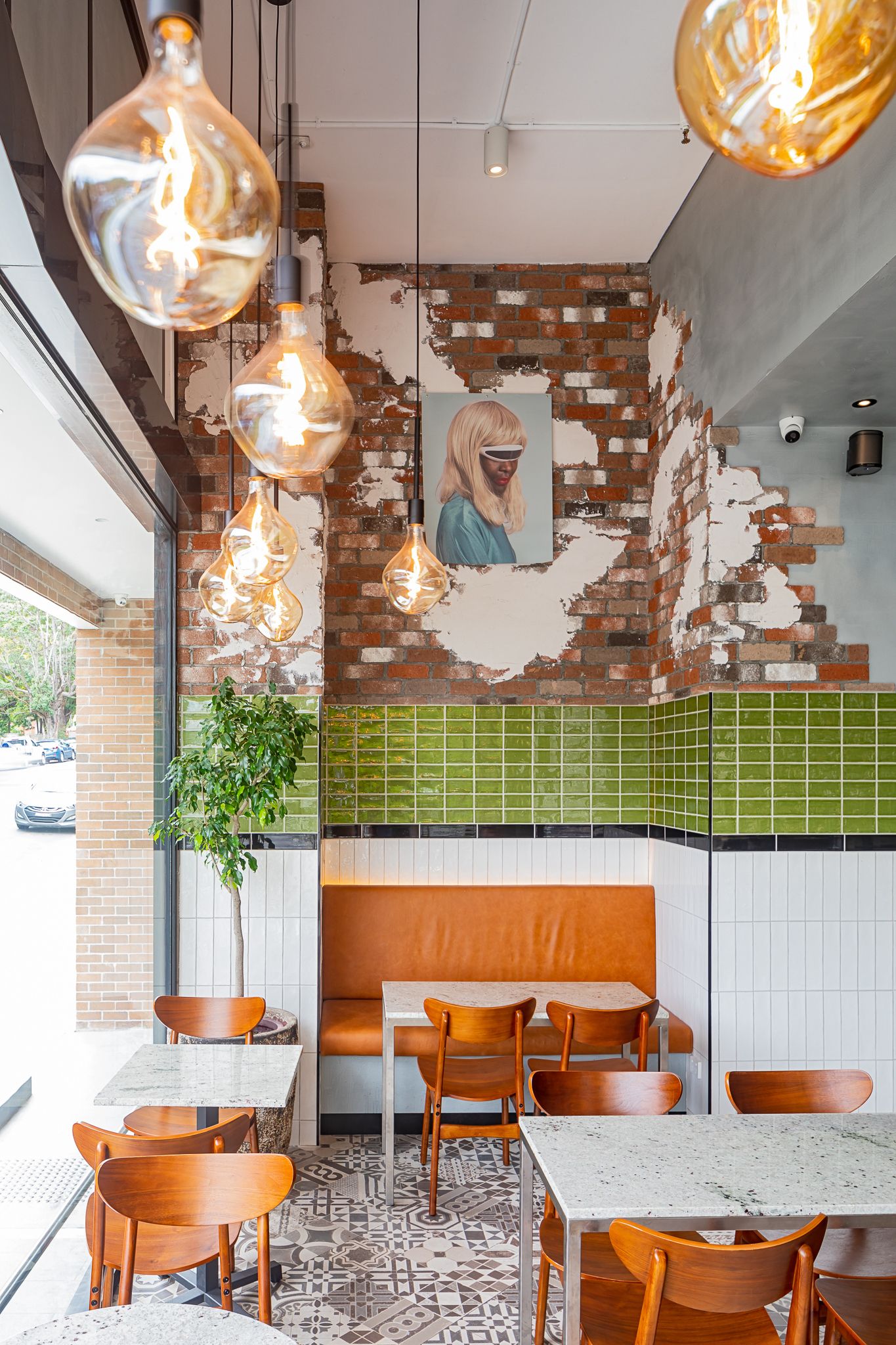 Stylish cafe interior with exposed brick, green tiles, glass pendant lights, orange banquette seating and marble-top tables.
