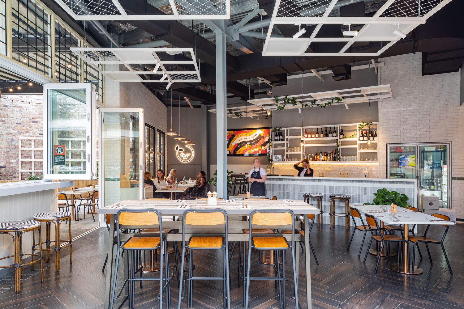 Modern restaurant interior with herringbone floors, wooden bar stools, communal tables and an open kitchen counter in the background.