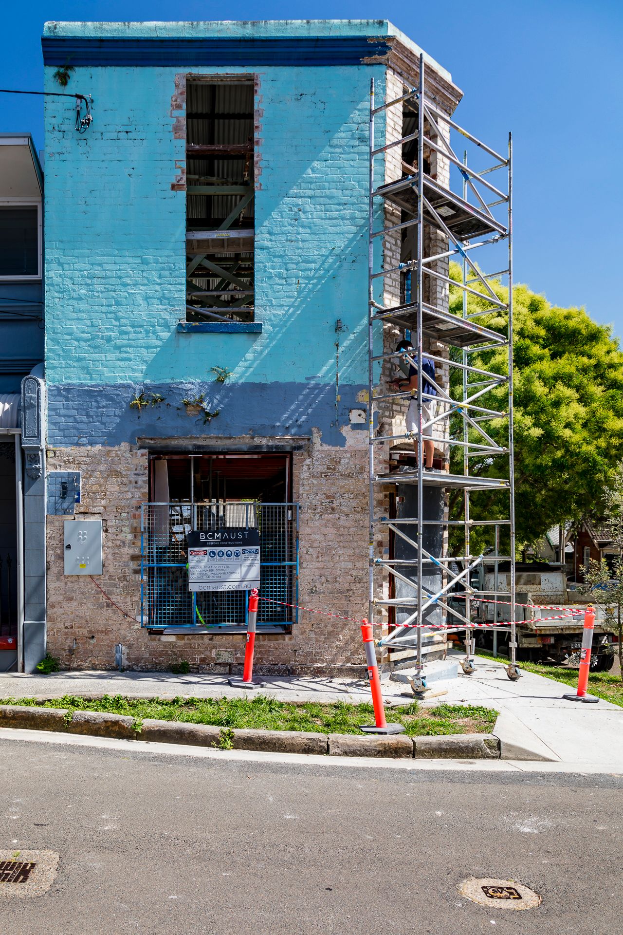 Blue-painted brick corner building under restoration with scaffold, safety cones and a contractor sign at street level.