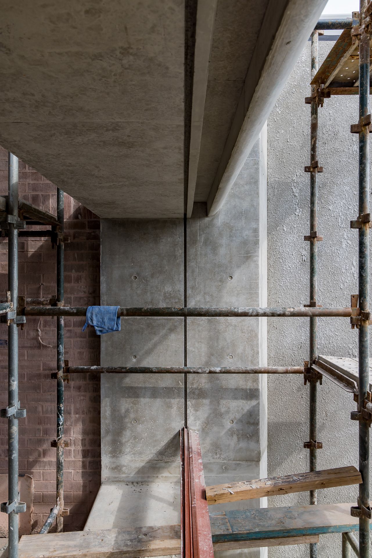 Scaffolding poles and a blue cloth in front of a raw concrete wall and red brick wall at an unfinished building site.