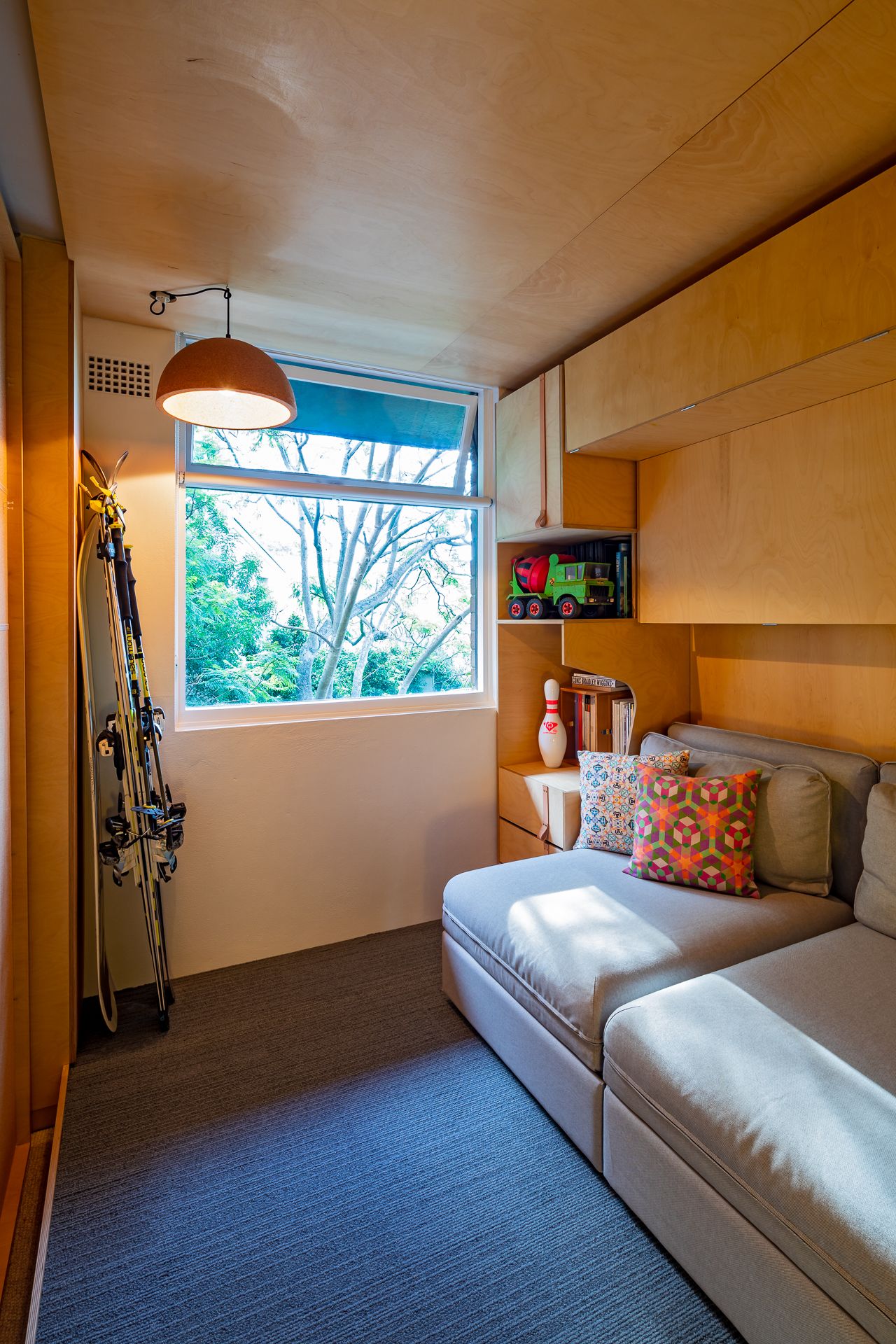 Compact plywood-clad bedroom with a built-in daybed, patterned cushions, pendant lamp, bright window and skis leaning against the wall.