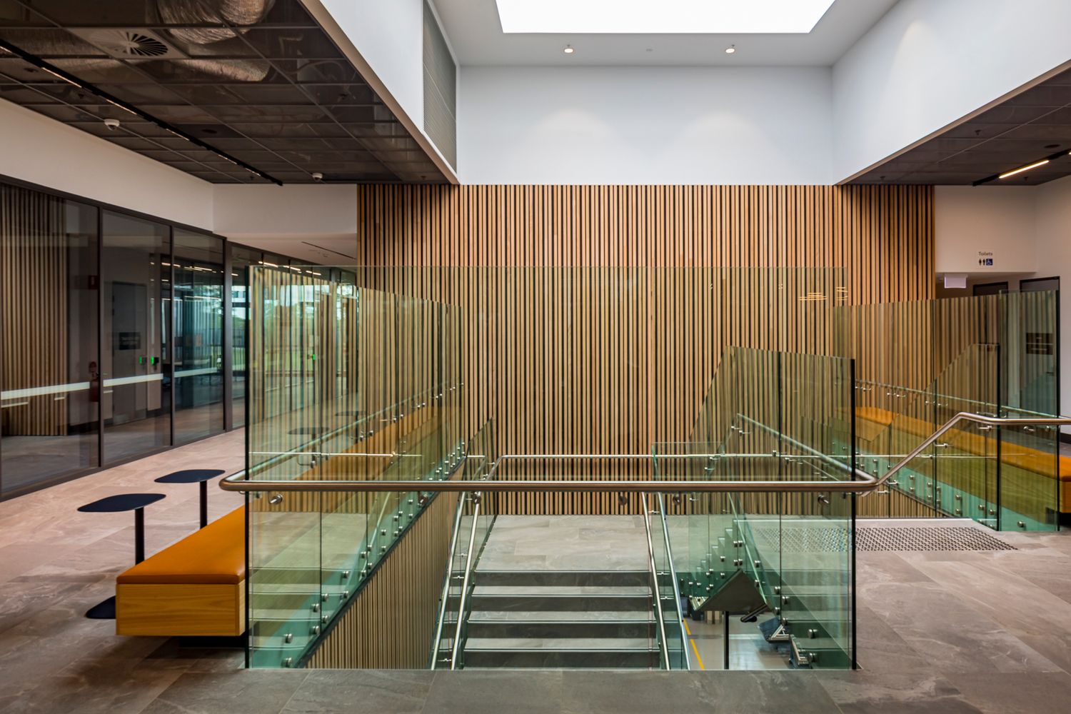 Modern campus foyer with glass-balustrade stairs, vertical timber wall cladding, and orange bench seating beside glazed meeting rooms.