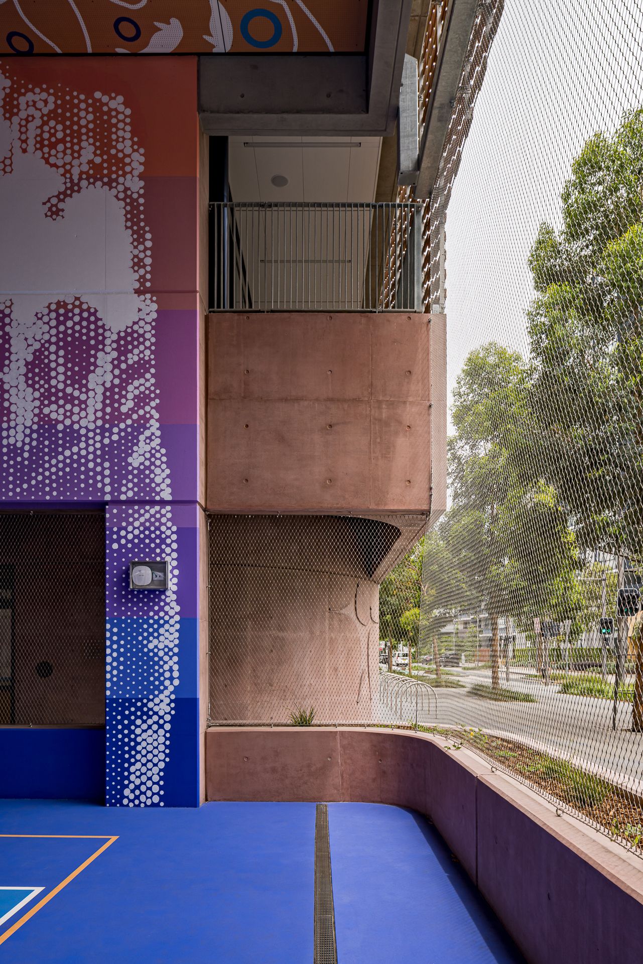 Blue sports court corner with a purple dotted mural, pink concrete walls and mesh fencing overlooking a tree-lined street.