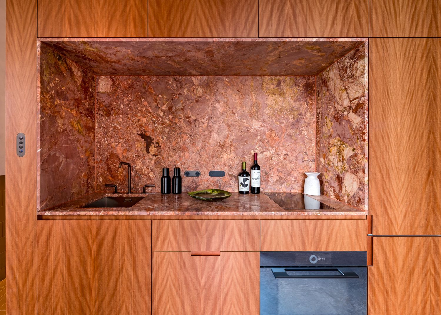 Timber cabinetry kitchenette with a recessed rose-toned marble alcove, small sink, built-in oven and wine bottles on the counter.