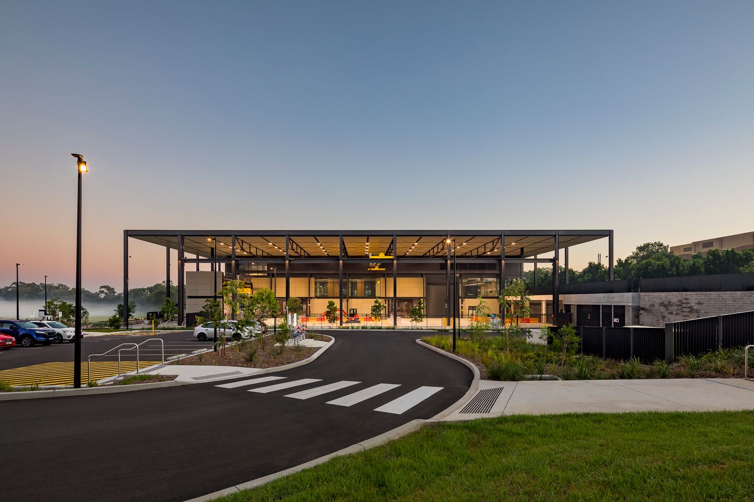 Contemporary steel-framed pavilion building glowing at dusk with a curved driveway, crosswalk and streetlight in the foreground.