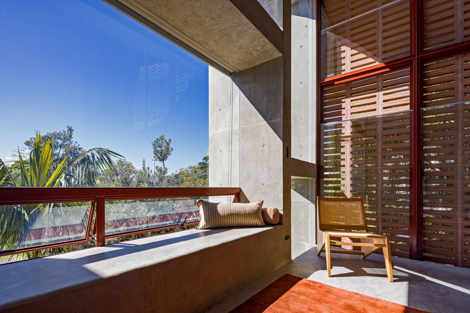 Sunlit concrete balcony with a built-in bench, cushion, timber chair and louvred screens facing palm trees and blue sky.