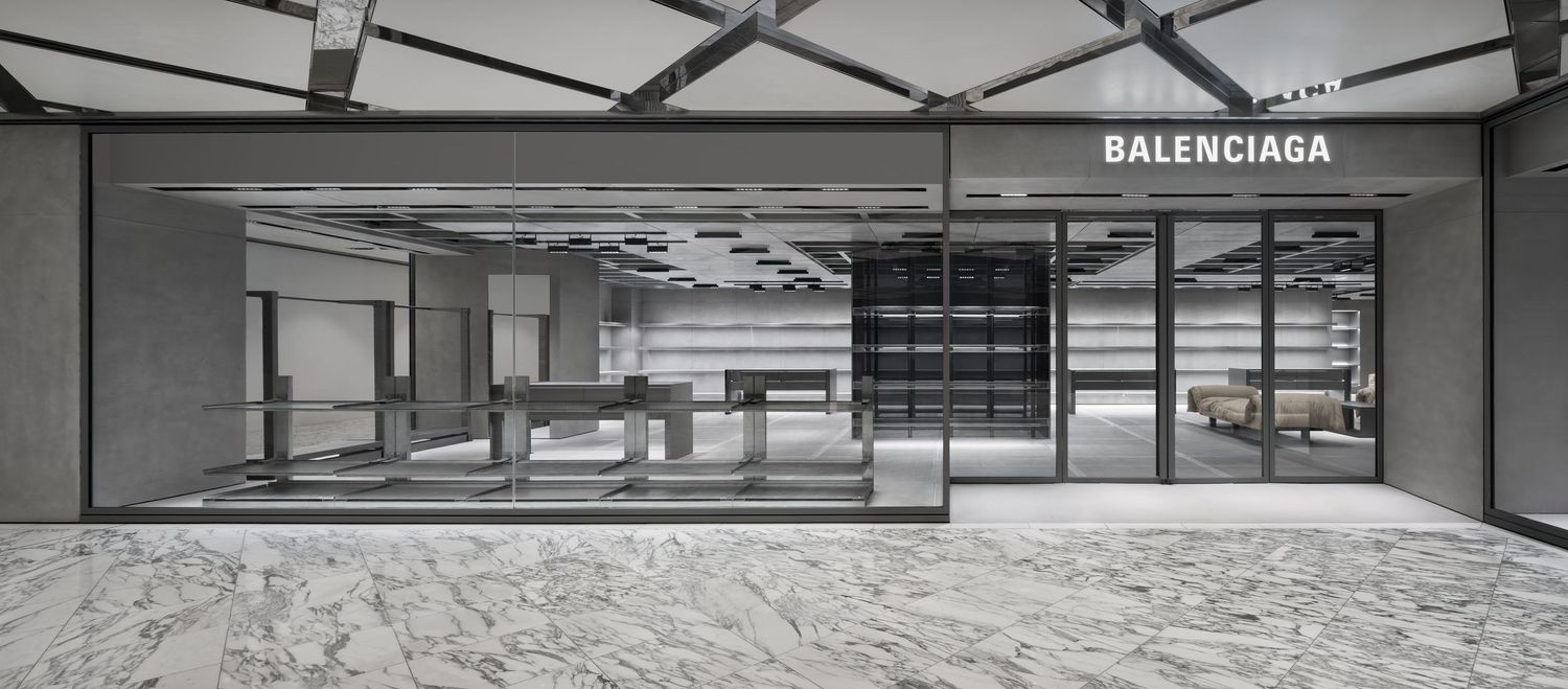 Entrance to a minimalist Balenciaga boutique with a glass storefront, dark interior shelving, and polished marble flooring.