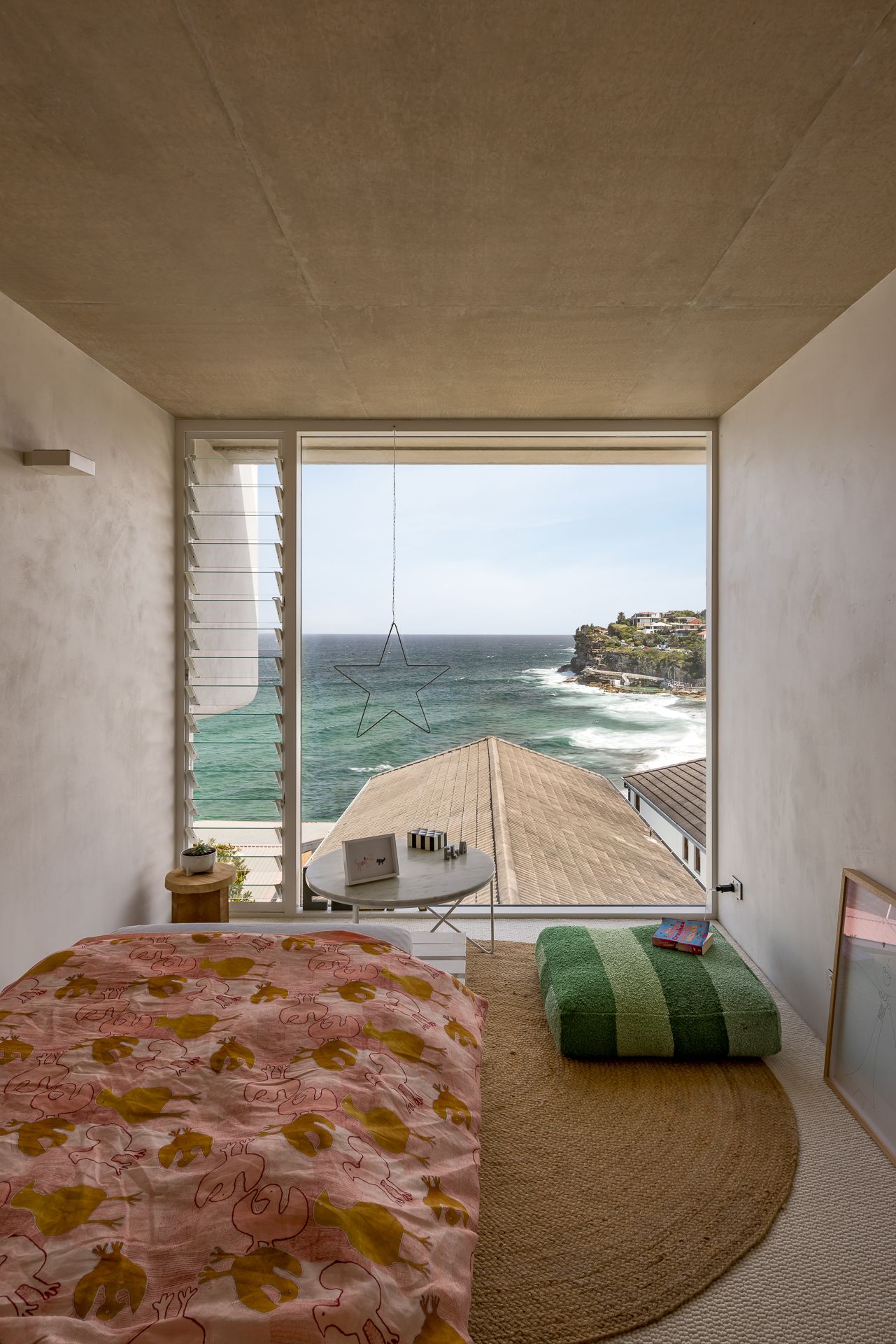 Minimalist bedroom with a pink floral quilt, round jute rug and a large picture window overlooking turquoise sea and coastal cliffs.