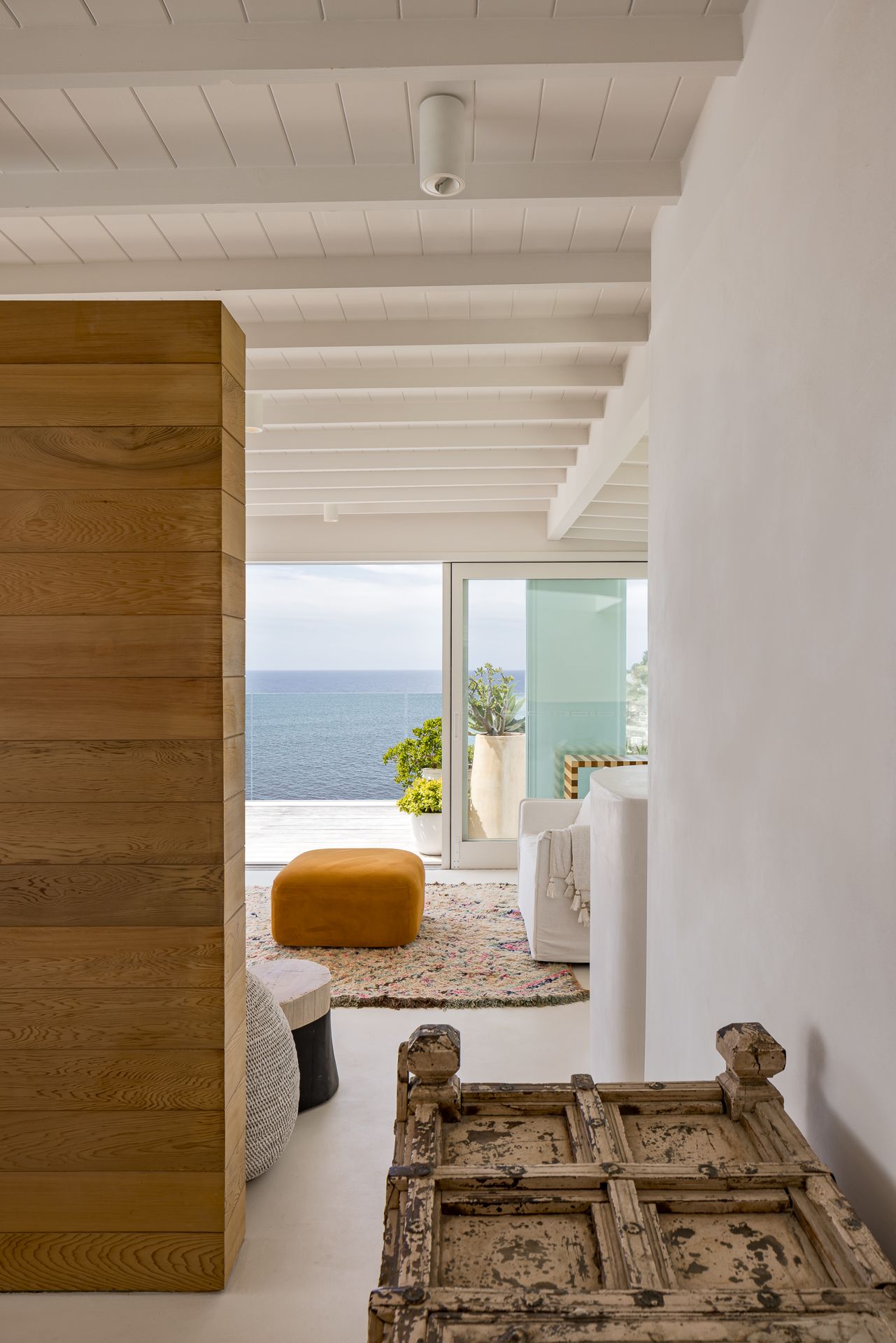 Sea view through a coastal home interior with timber wall panel, mustard ottoman, Moroccan rug and vintage carved wooden chest.