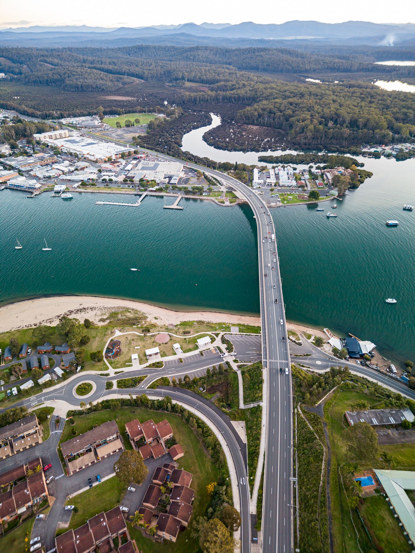 Aerial view of a long road bridge crossing a wide blue estuary beside a sandy beach, waterfront homes and green hills.