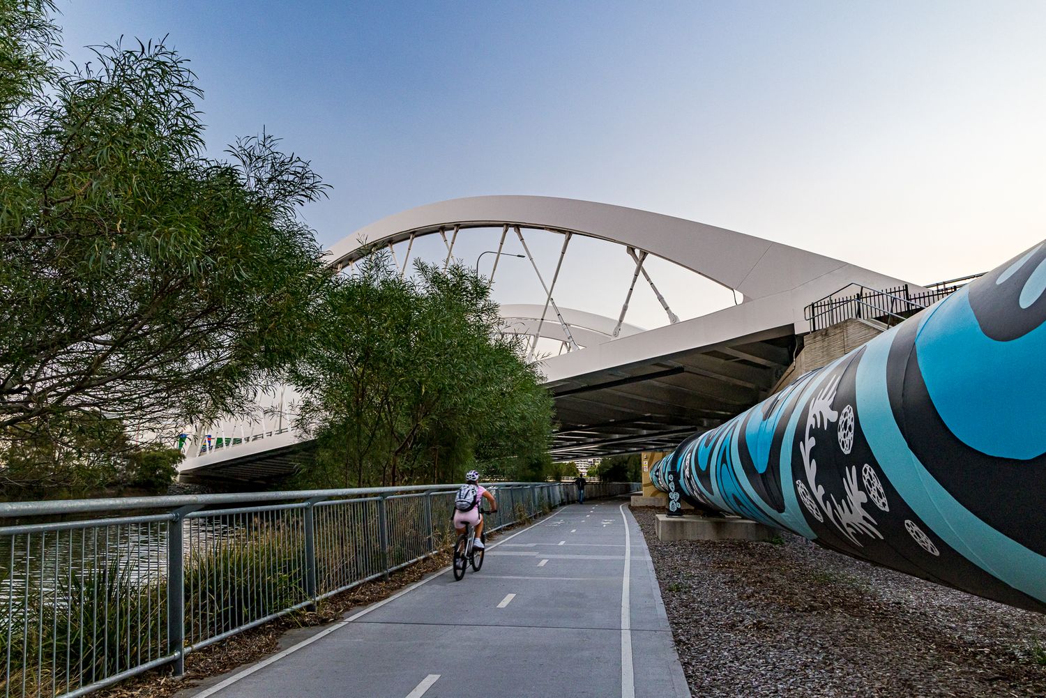 Cyclist rides along a riverside path beneath a white arch bridge, beside a blue and black graphic-wrapped pipe.