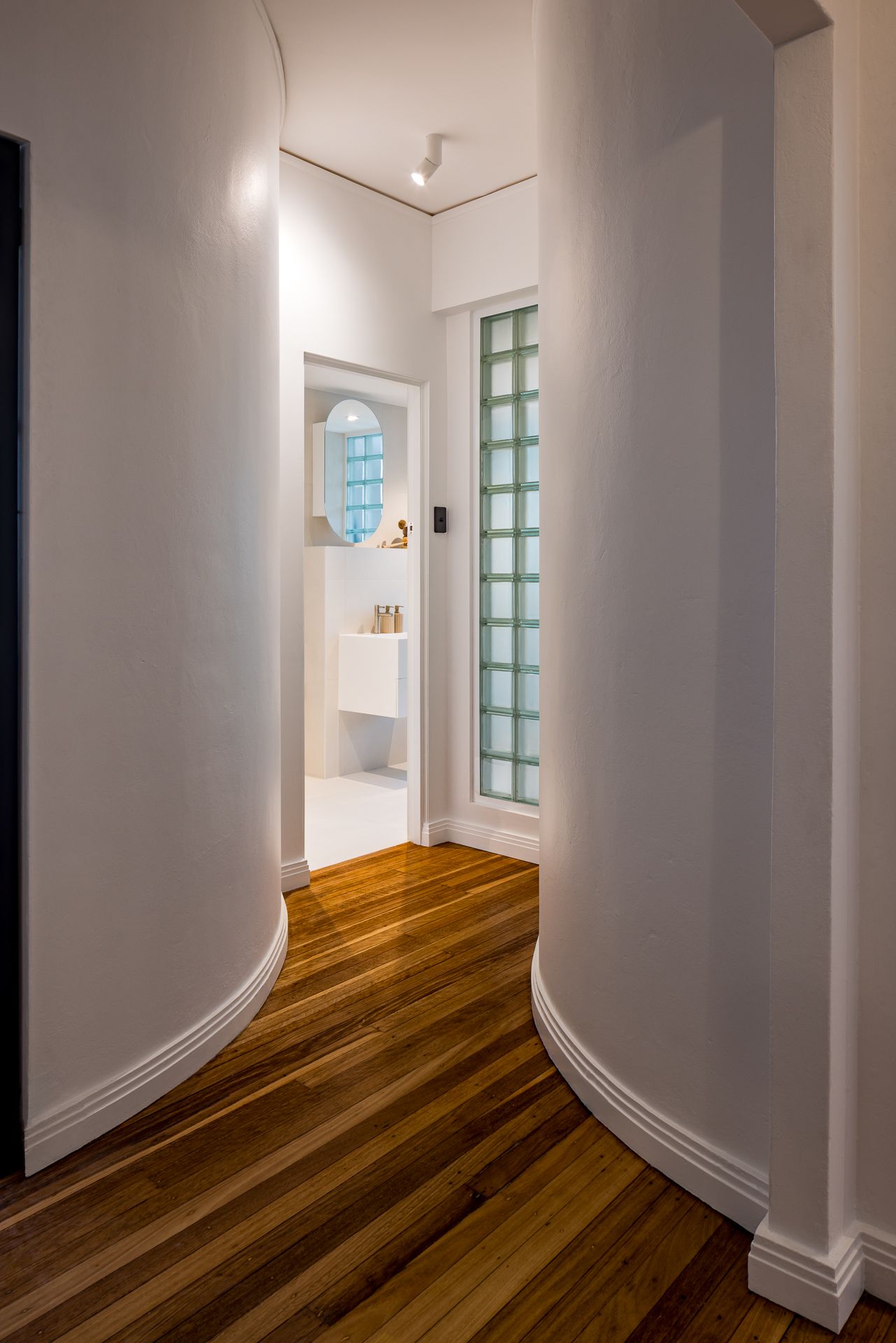 Curved white hallway with polished timber floors leads to a bathroom with a green-tinted glass block window.