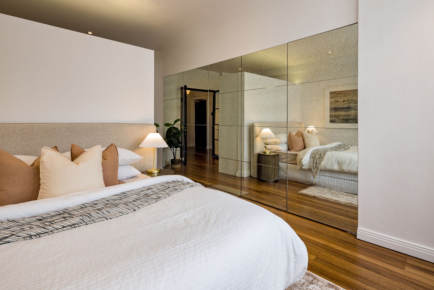 Spacious bedroom with a upholstered headboard, neutral cushions, mirrored wardrobe doors and warm polished timber flooring.