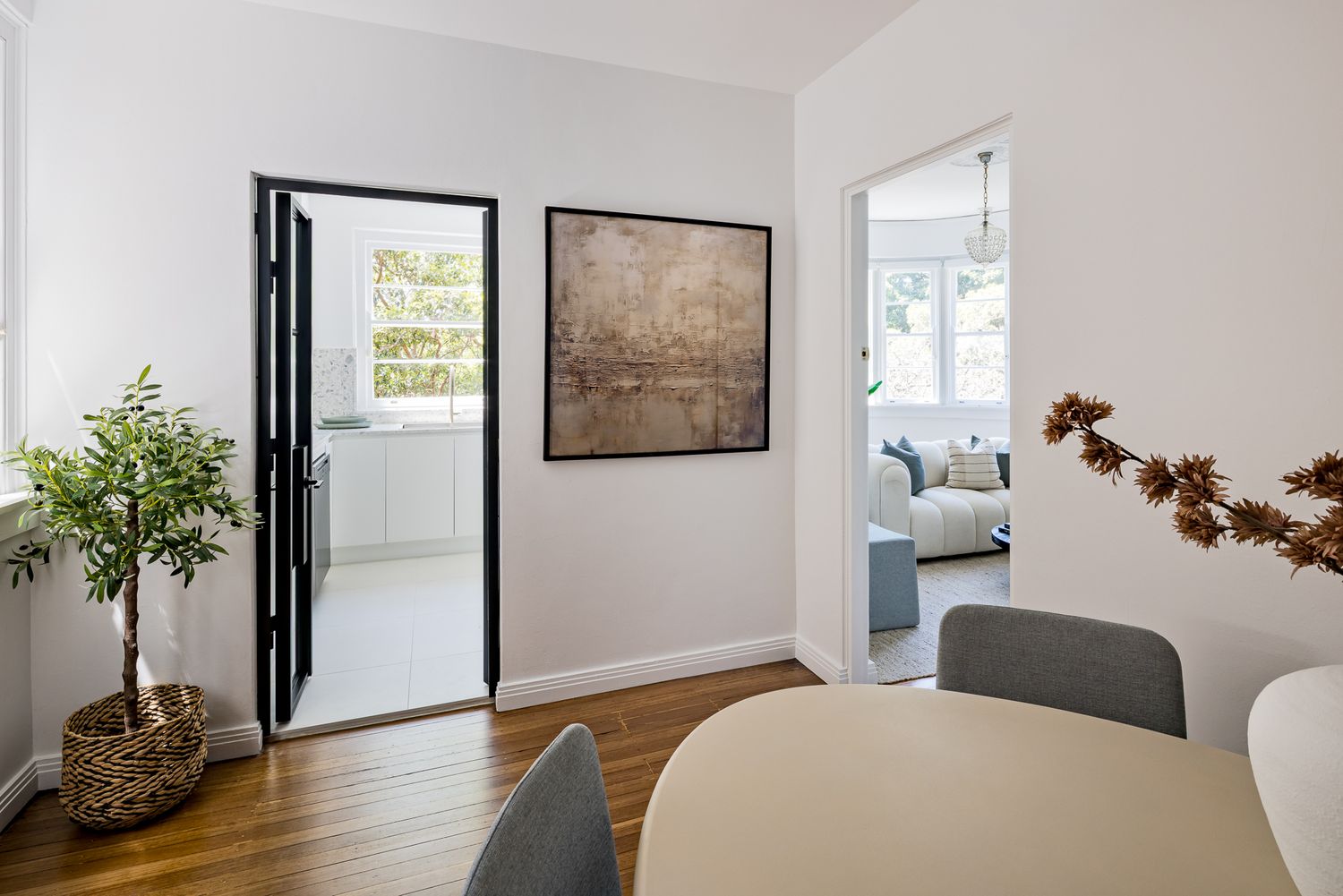 View from dining area through black steel-framed doorway into a bright white kitchen, with potted plant and framed artwork.