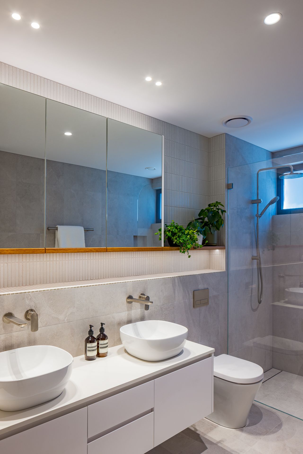 Modern bathroom with double white vessel sinks, floating vanity, mirrored cabinets, walk-in glass shower and wall-hung toilet.