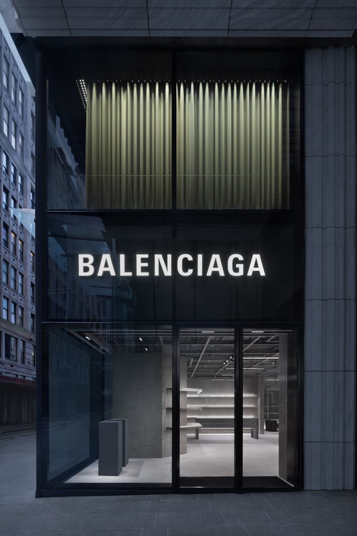Balenciaga flagship store exterior at dusk with illuminated white logo, glass facade and minimalist grey interior visible inside.