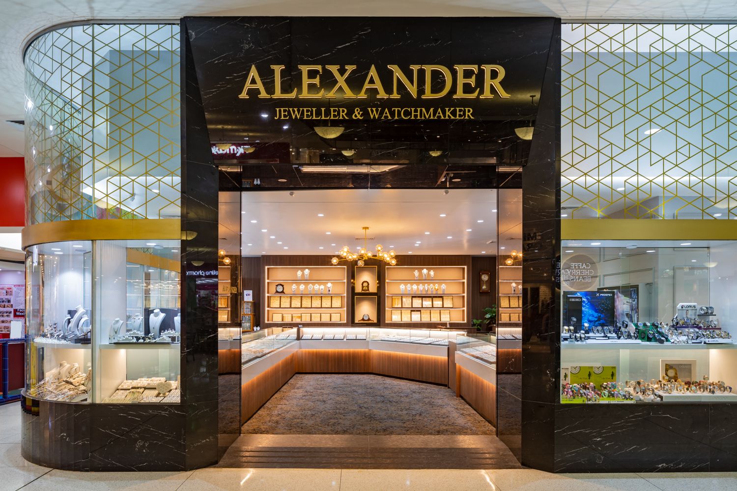 Alexander Jeweller & Watchmaker boutique entrance with gold lettering, lit display cabinets and patterned glass in a shopping centre.