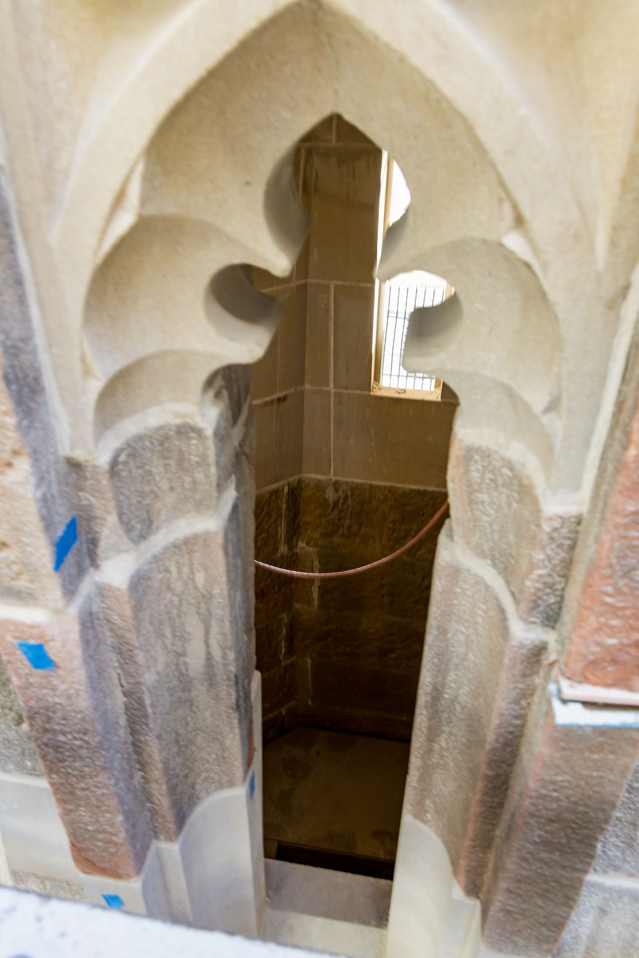 Looking down through a Gothic stone window opening with trefoil tracery into a shaft with a rope and blue tape markers.