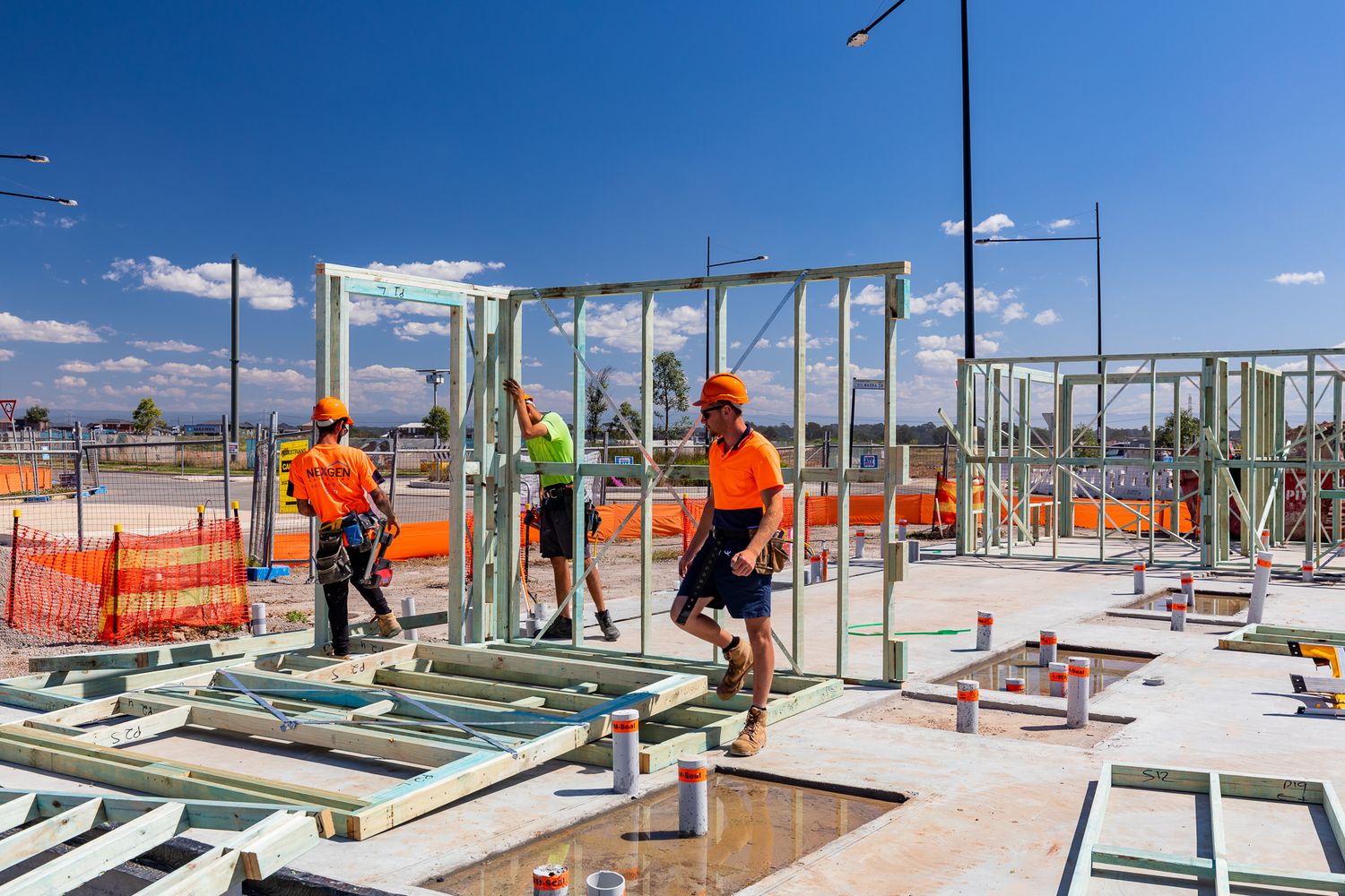Construction workers in orange hi-vis shirts and hard hats assembling green steel wall frames on a concrete slab at a housing site.