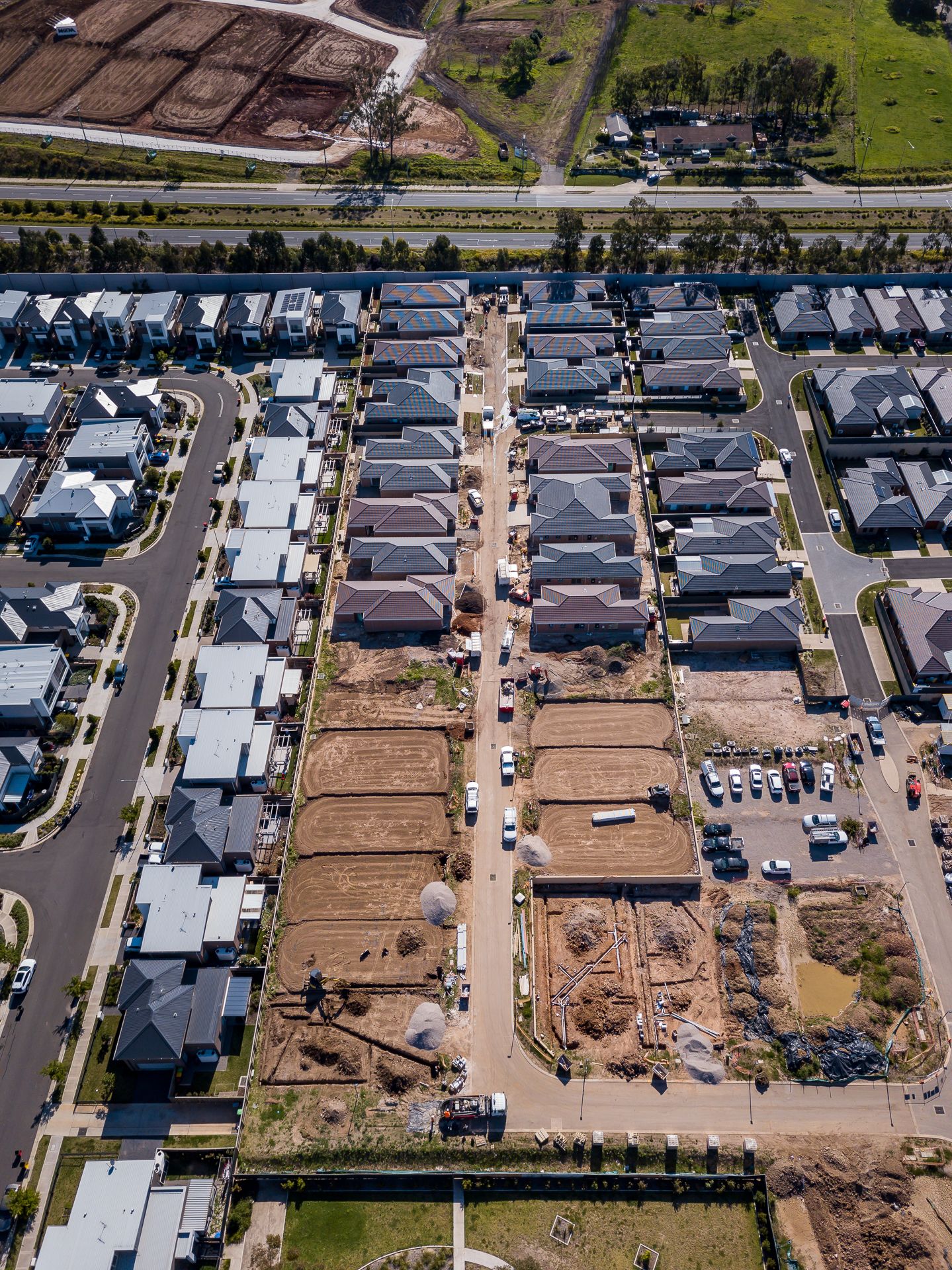 Overhead drone view of a new housing estate with finished townhouses, dirt building lots and streets lined with parked cars.