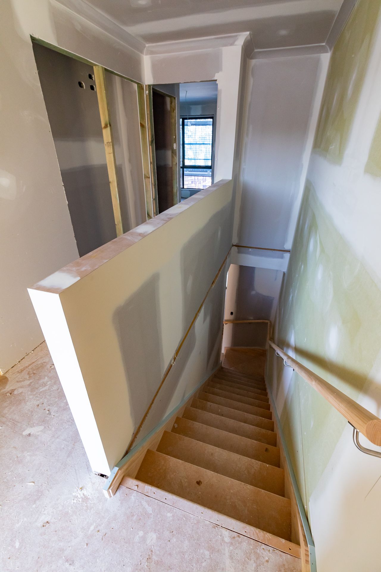 Unfinished staircase with bare timber treads, plasterboard walls and a handrail leading down in a home under construction.