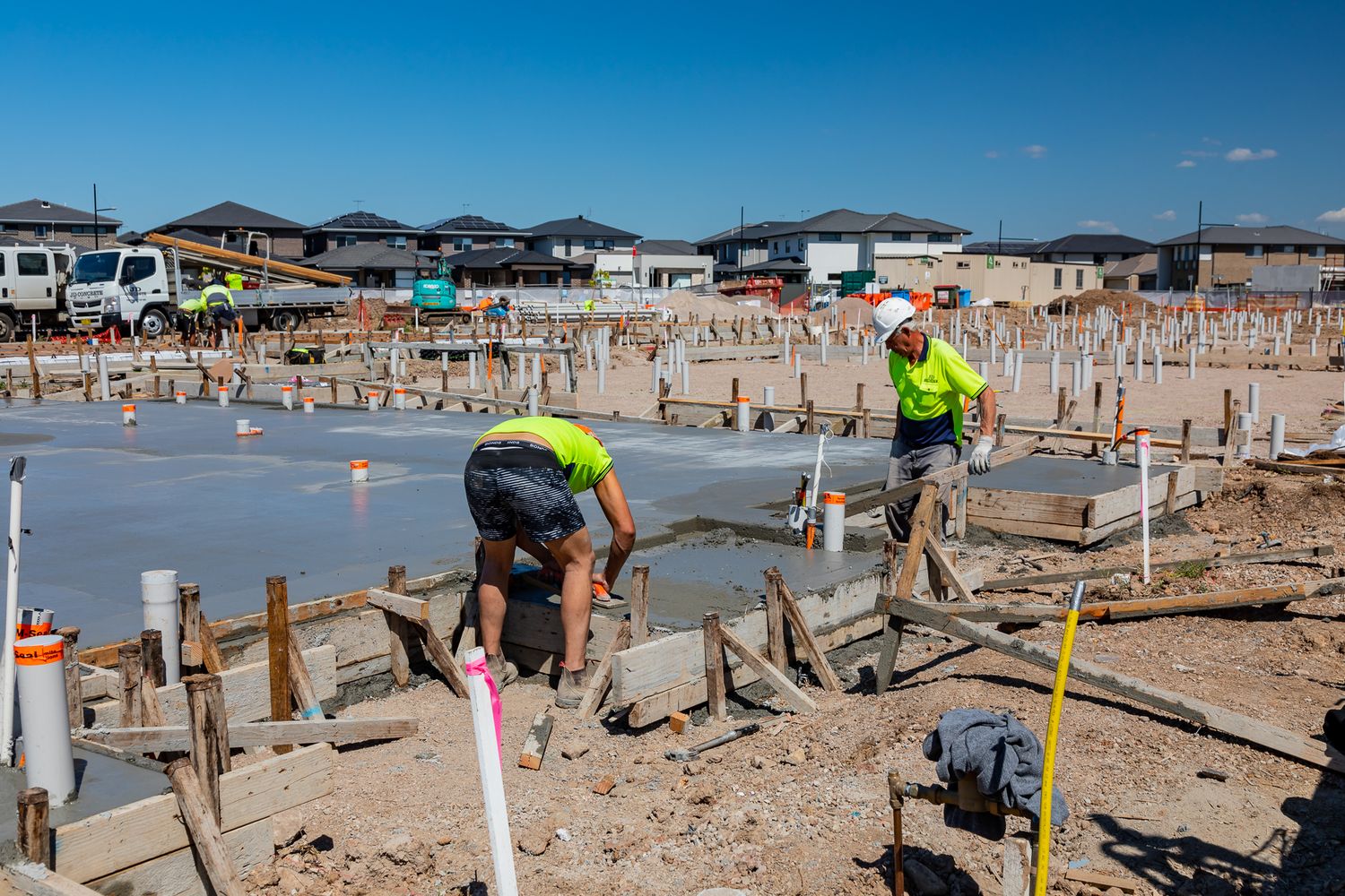 Construction workers in hi-vis shirts pour and level a concrete slab inside timber formwork at a large housing development site.