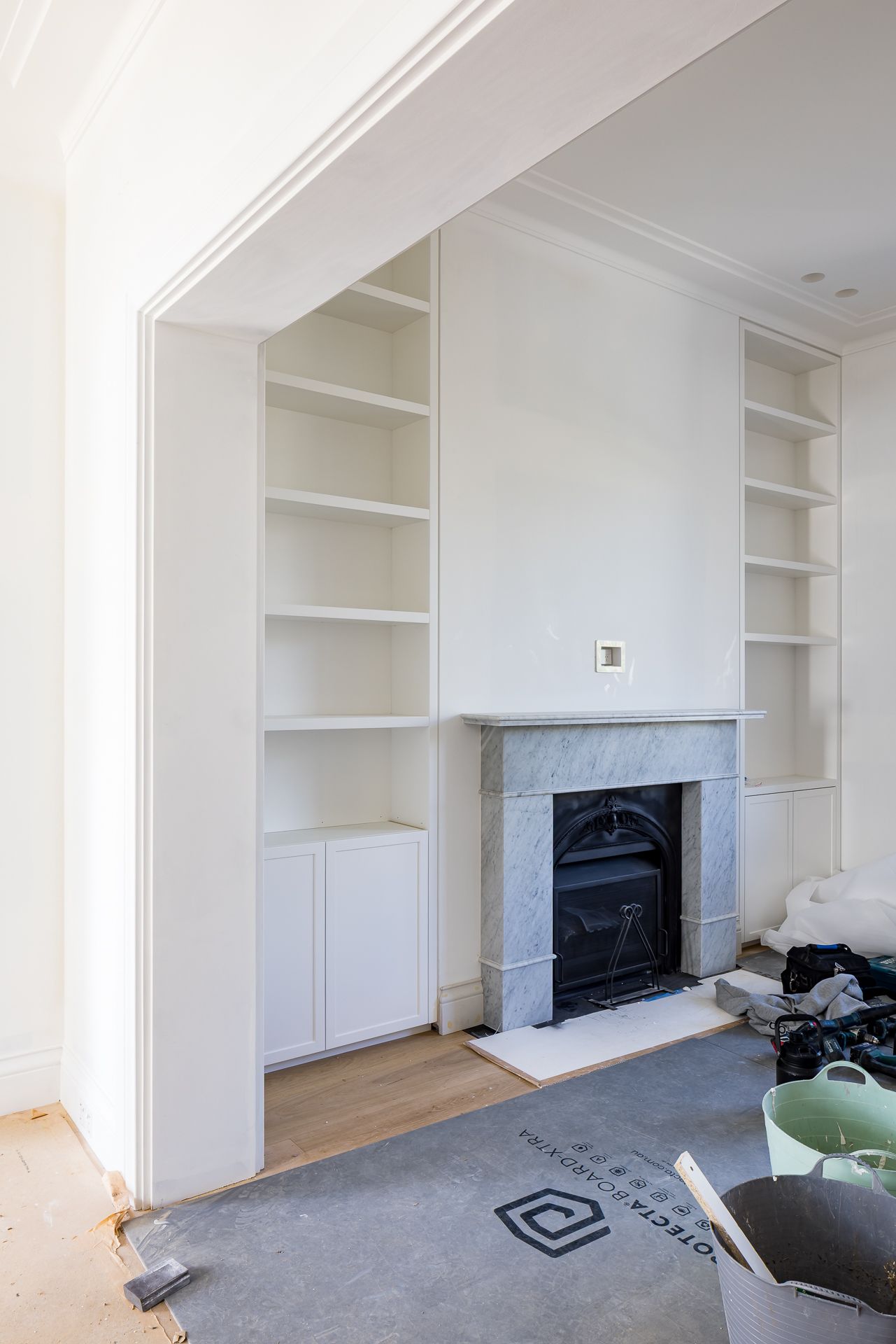 White built-in bookshelves flanking a marble fireplace in a renovated living room with timber flooring and protective floor covering.
