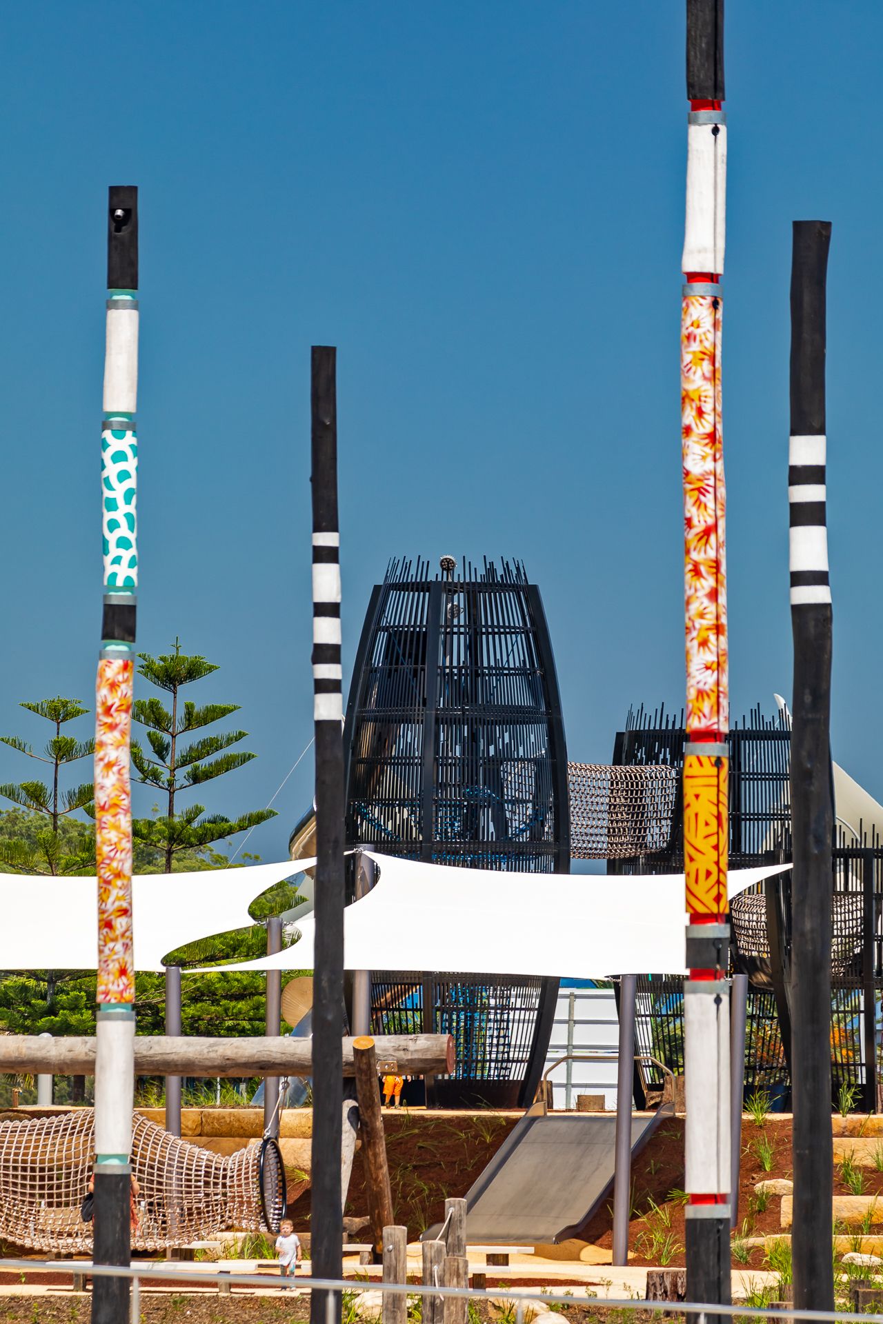 Tall painted Aboriginal-style poles stand beside white shade sails and a black lattice tower at a modern playground.