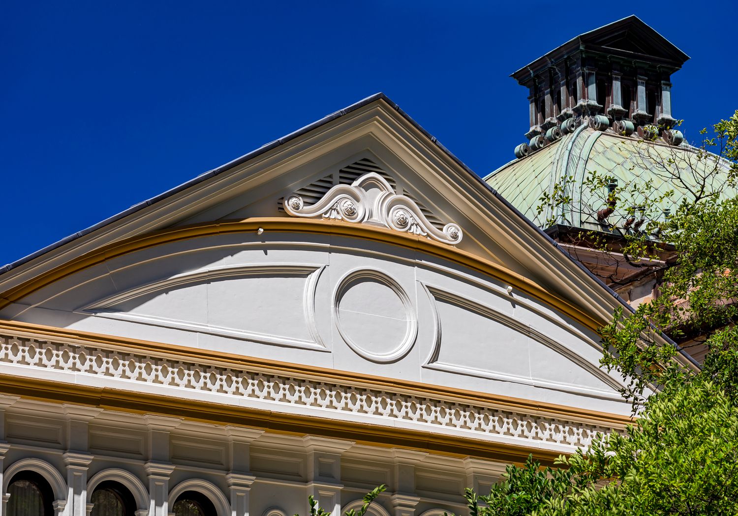 Detail of a heritage building pediment with ornate plaster scrollwork, circular medallion and decorative cornice beside a green copper dome.