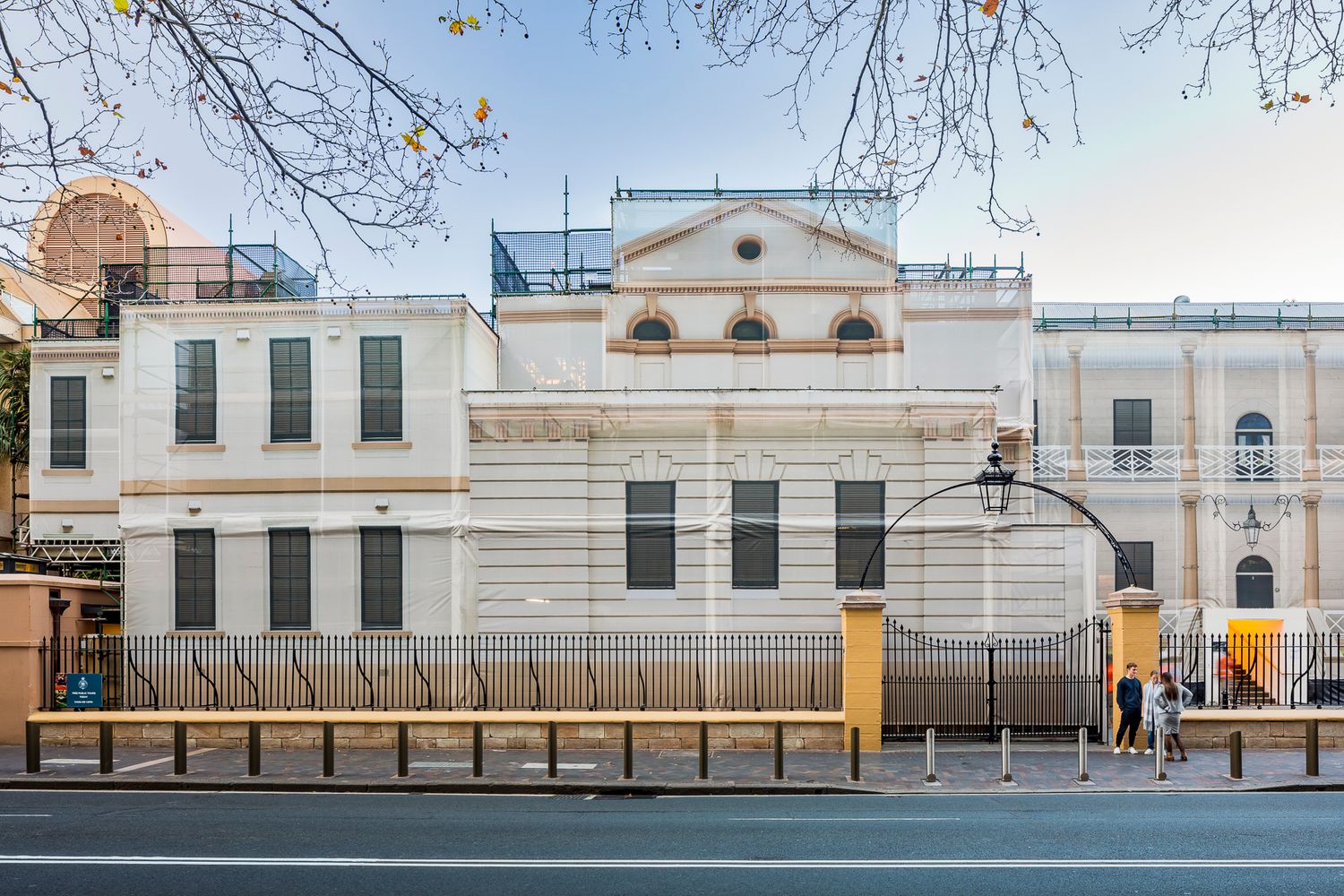 Historic white building with arched windows and scaffolding on the roof, viewed from the street through bare tree branches.