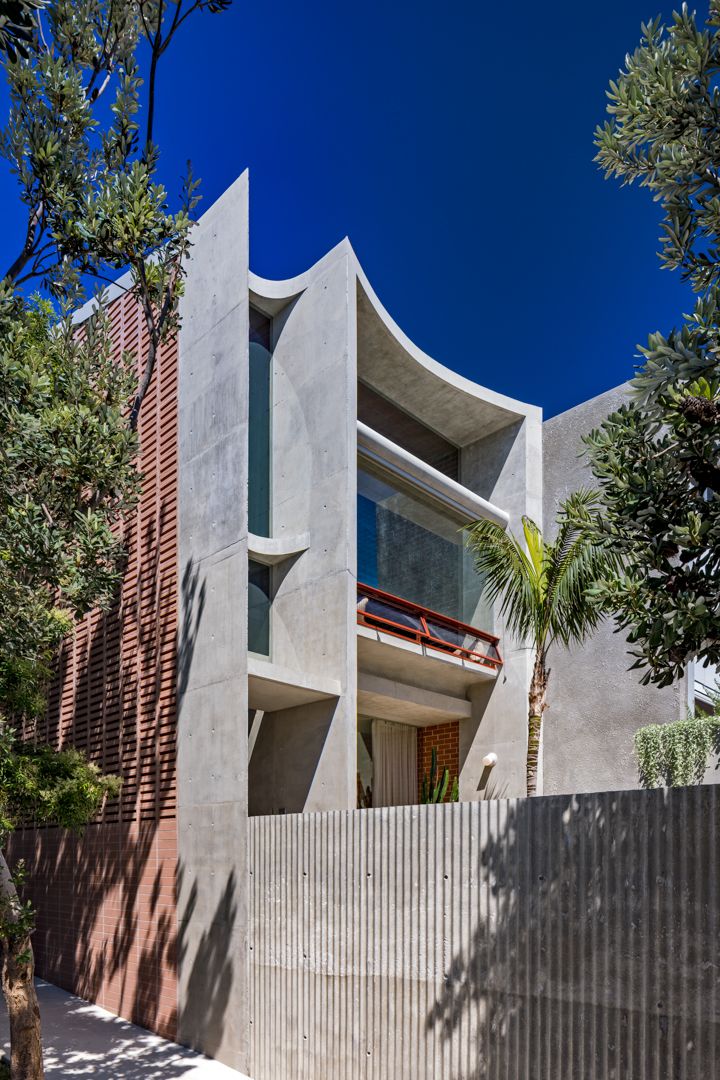 Sculptural concrete townhouse facade with curved forms, a brick screen wall, a red balcony railing and palm foliage.