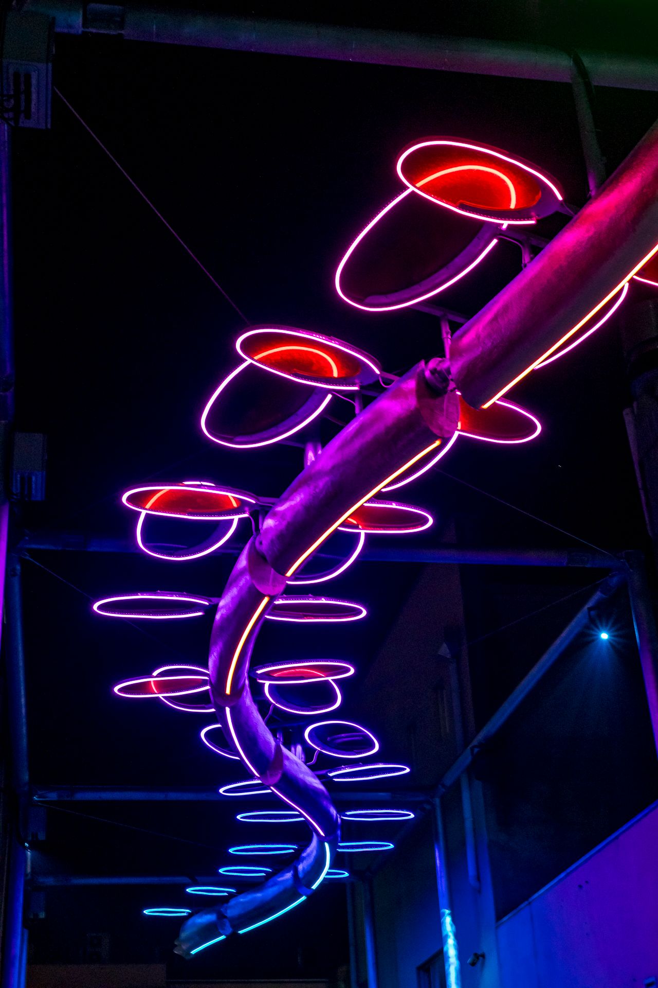Close-up of a glowing neon sculpture with pink, purple and orange rings spiraling around a metallic tube against a dark night sky.