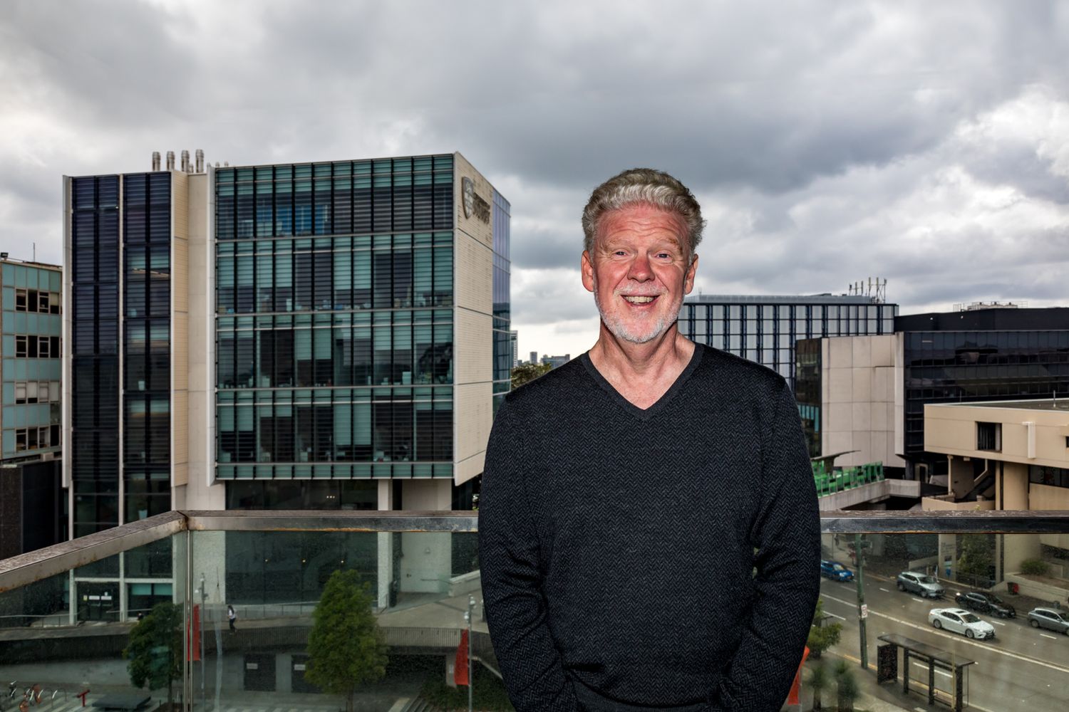 A smiling gray-haired man in a dark sweater stands on a balcony with modern glass office buildings and a cloudy sky behind him.