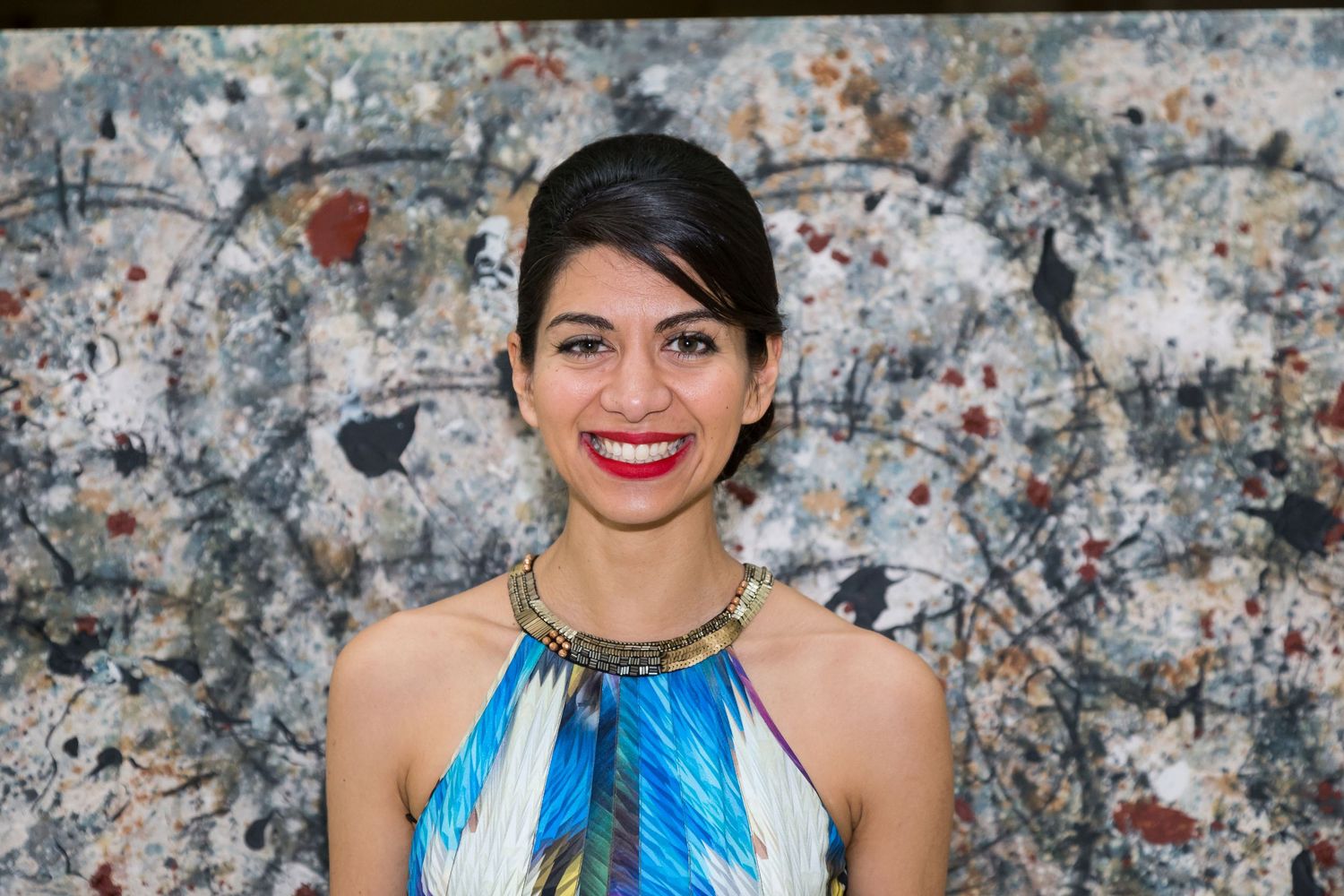 Smiling woman with dark hair and red lipstick wears a blue patterned top and gold necklace in front of an abstract splatter painting.