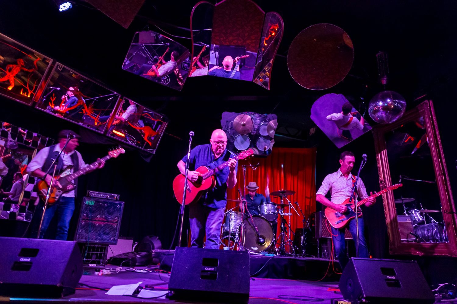 Three musicians play guitars on a dimly lit stage with a drum kit behind them, surrounded by amplifiers and colorful stage lighting.