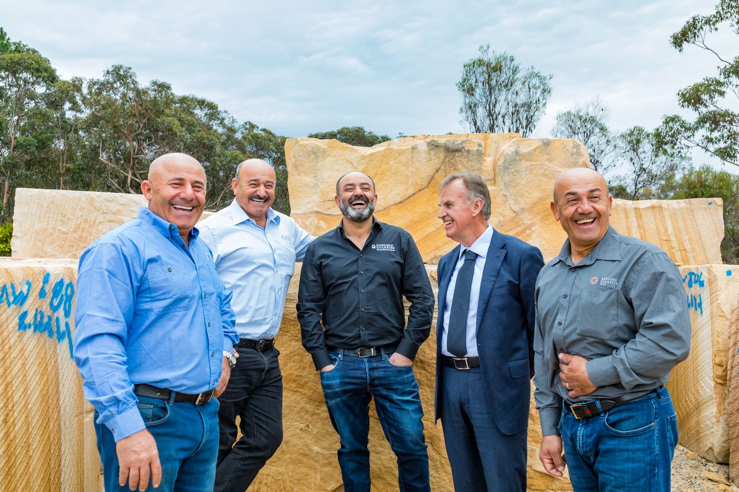 Five smiling men stand together outdoors in front of large sandstone blocks at a quarry, dressed in casual and business attire.