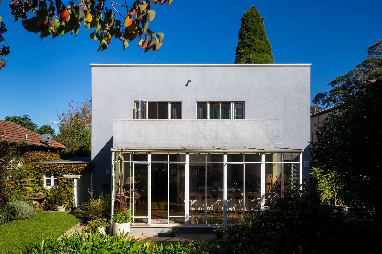 Modernist white rendered house with a glass sunroom and rectangular windows, surrounded by lush garden greenery and trees.