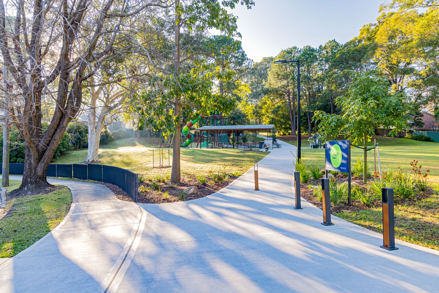 Sunlit concrete pathways with bollards lead through a green park toward a covered picnic shelter surrounded by mature trees.