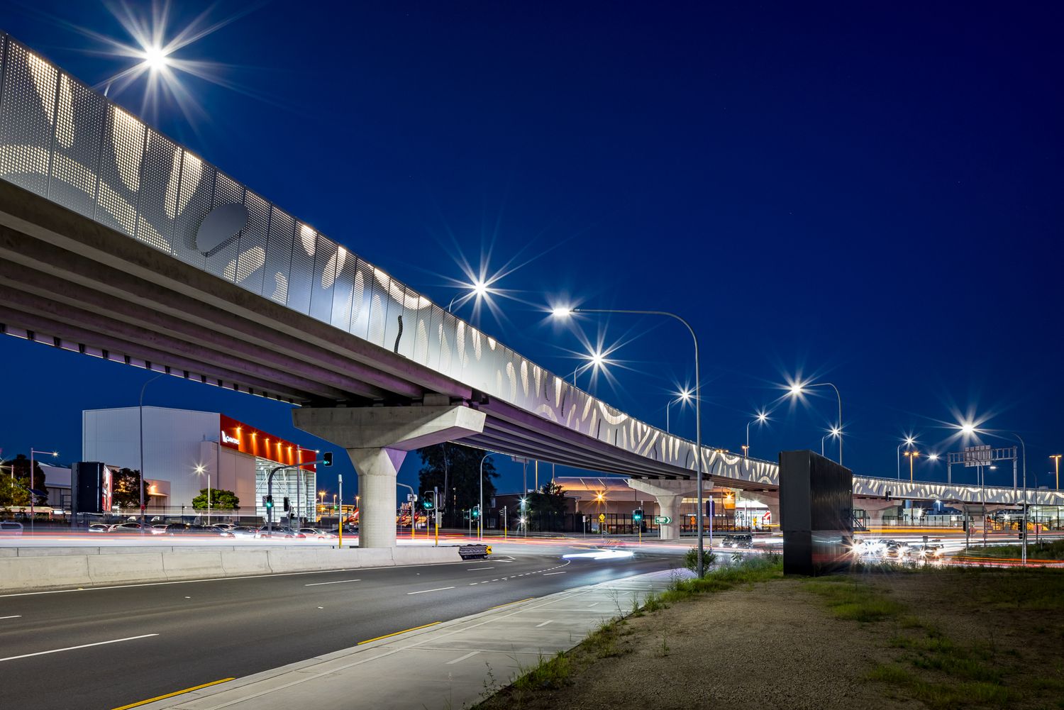 Elevated rail viaduct with a patterned perforated metal fascia curves over a road at dusk beneath a deep blue sky and bright street lights.